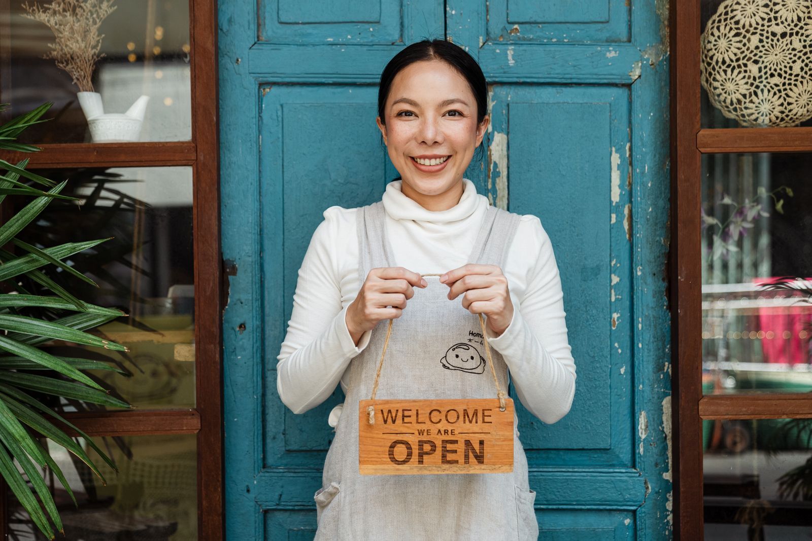 Dona de uma cafeteria sorrindo com avental, mostrando um cartaz de boas-vindas perto de uma porta azul