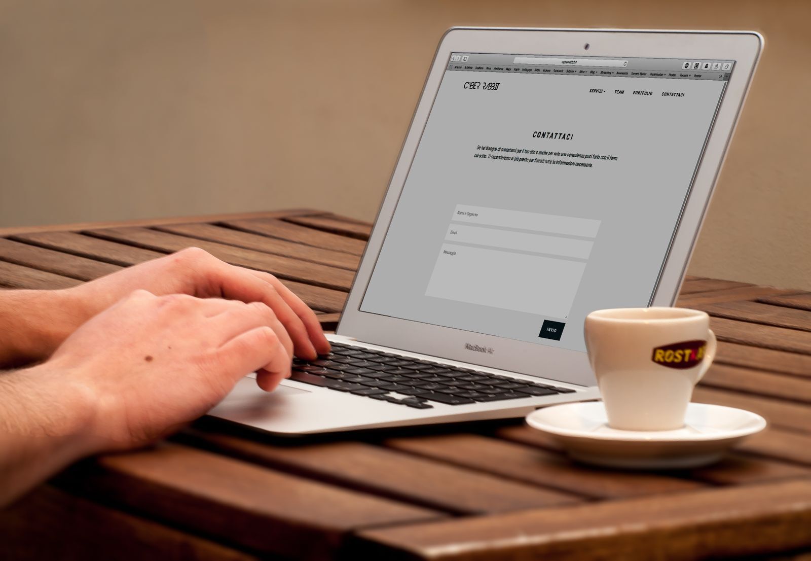 Mãos digitando em um notebook ao lado de uma xícara sobre uma mesa de madeira